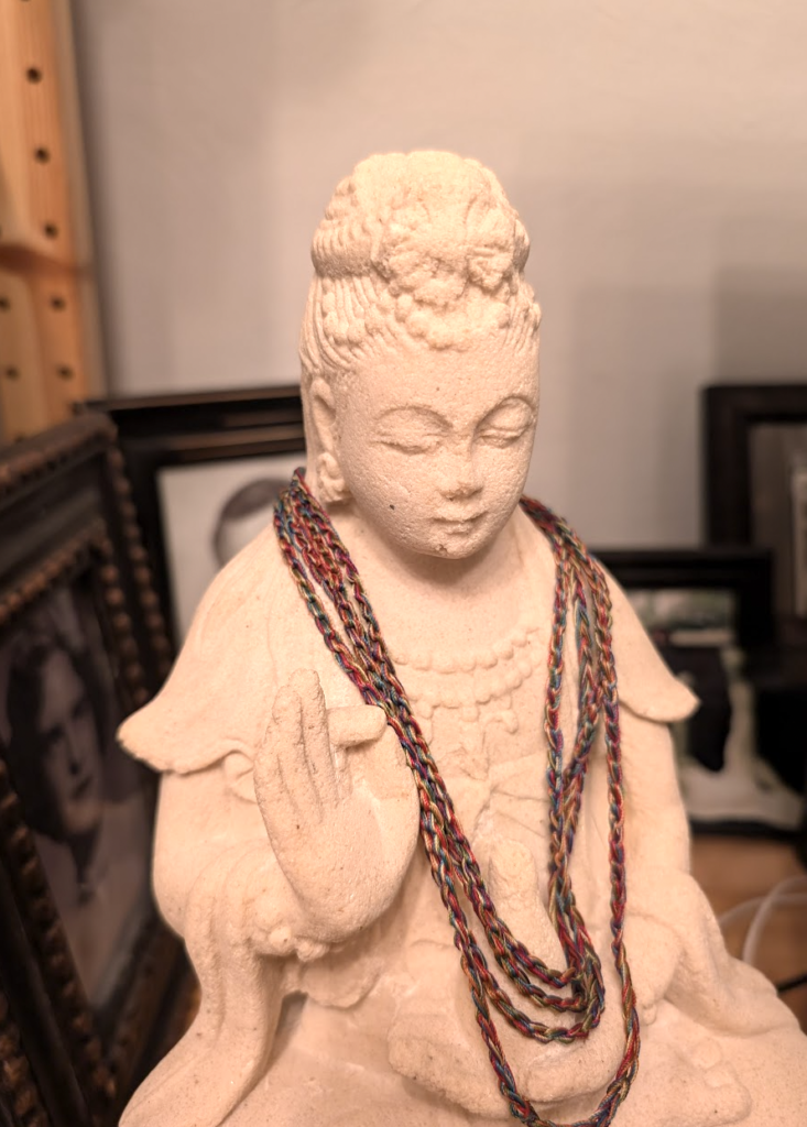 A picture of a white stone statue of the Boddhisatva Avalokiteshvara with a colorful lanyard hanging around the neck. Avalokiteshvara has their eyes closed, and is holding their hand with forefinger and thumb pinched, while the other cradles a bottle or carafe.
